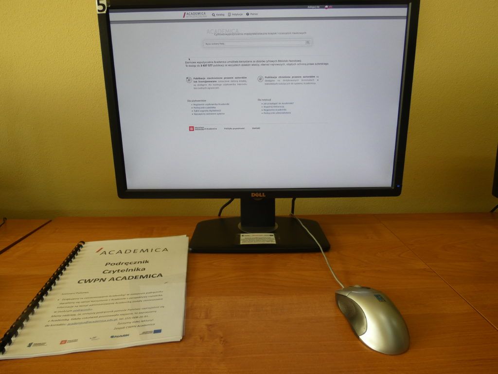 Na zdjęciu biurko na którym stoi komputer z uruchomioną przeglądarką academici na biurku leży po lewej stronie Podręcznik obsługi academici