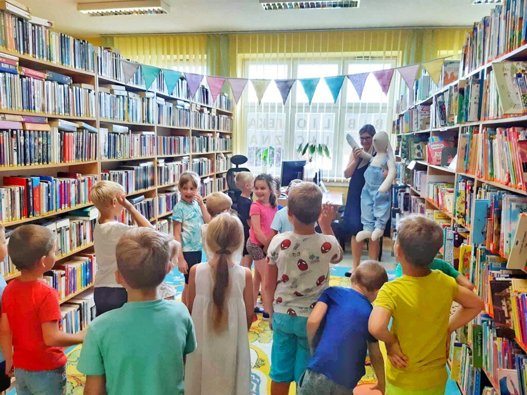 Grupa dzieci stoi w bibliotece, zwrócona do bibliotekarki, która trzyma ogromną maskotkę Królika