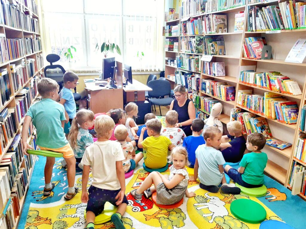 Grupa dzieci siedzi na kolorowym dywanie w bibliotece, kobieta czytająca dzieciom książkę