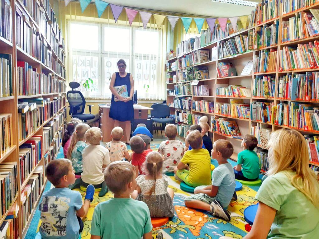 Grupa dzieci siedzi na dywanie, bibliotekarka stoi mówiąc do dzieci