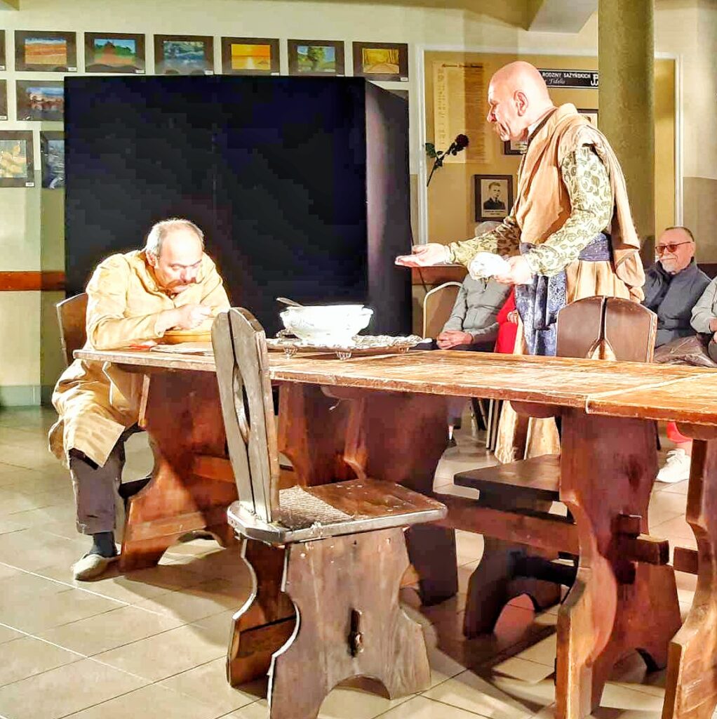 Scena ze spektaklu "Zemsta". Cześnik siedzi przy stole i je zupę, Śmigielski  stoi przy stole i coś tłumaczy Cześnikowi