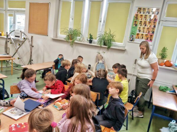duża grupa dzieci siedzi przy stolikach i ogląda książki, razem z dziećmi siedzi duża maskotka królik, po prawej stronie stoi nauczycielka, która tłumaczy zadanie do wykonania